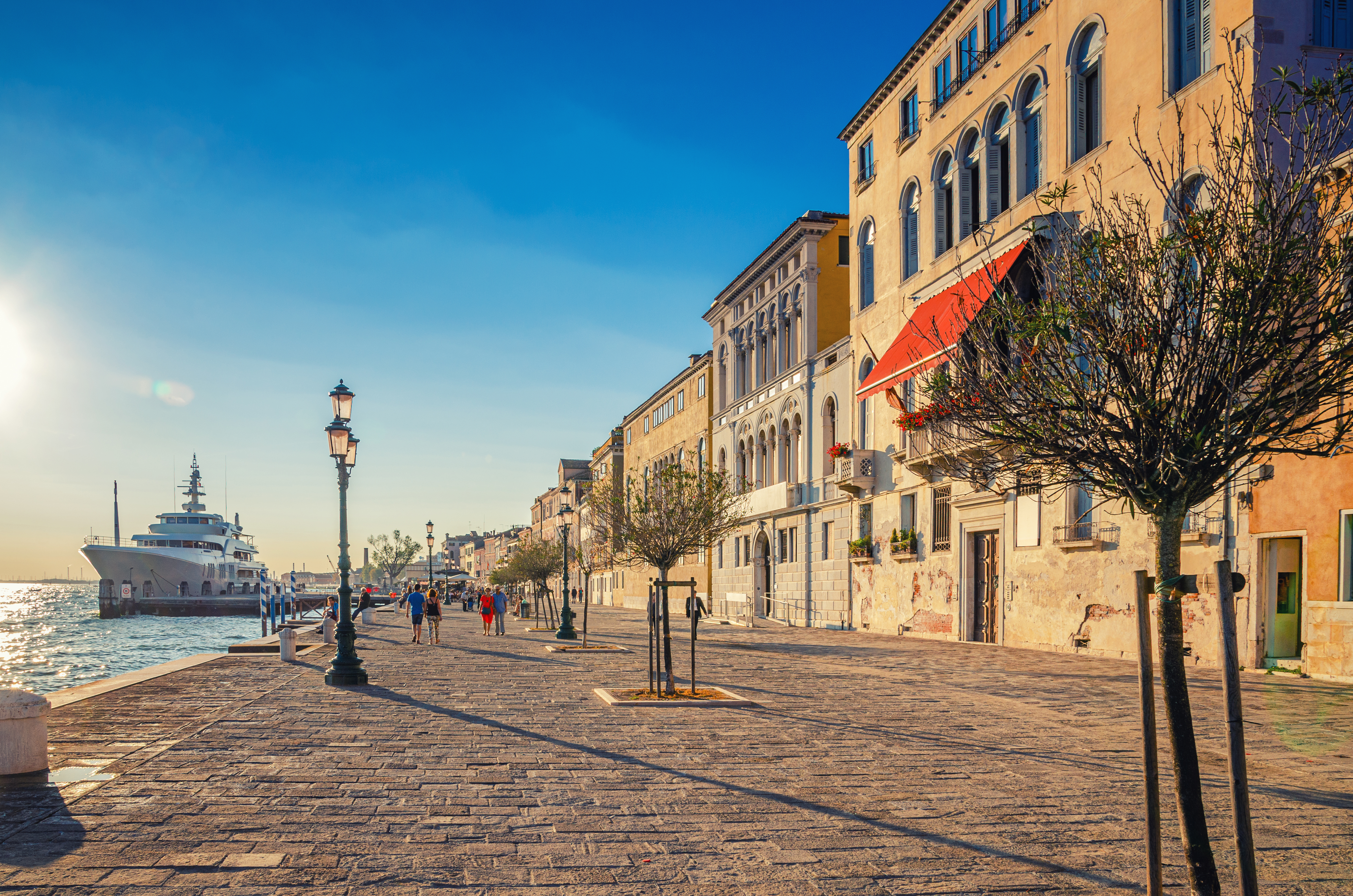 People tourists are walking down embankment promenade Fondamenta Zattere Al Ponte Lungo with trees and yacht moored in water of Giudecca canal waterfront in Venice